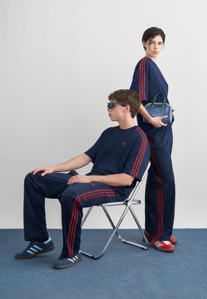 Άνδρας καθισμένος σε καρέκλα με σκούρο μπλε φόρμα με κόκκινες ρίγες και γυαλιά ηλίου· γυναίκα στέκεται δίπλα του με παρόμοιο σύνολο κρατώντας μια σκούρου μπλε τσάντα Adidas.