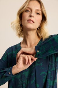 Kobieta z krótkimi blond włosami, nosząca niebieską i zieloną kwiecistą bluzkę z długim rękawem, poprawiająca mankiet swojej koszuli z metalowym detalem guzikowym.