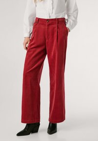 Pantaloni larghi in corduroy rosso con texture, pieghe frontali, tasche laterali e chiusura a bottone, abbinati a stivaletti neri con tacco.