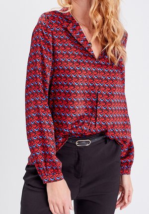 Femme portant un chemisier rouge à manches longues avec un motif géométrique bleu et noir, rentré dans un pantalon noir avec une ceinture noire.