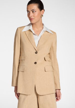 Vrouw die een beige, op maat gemaakte blazer draagt met twee knopen en vier zakken aan de voorkant, gecombineerd met een bijpassende broek met wijde pijpen en een wit overhemd met kraag.