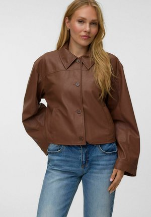 Femme aux longs cheveux blonds portant une veste en cuir marron boutonnée et un jean en denim bleu sur un fond clair uni.
