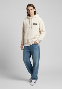 Felpa crema con la scritta "AMOUR" stampata in nero, dotata di coulisse e tasca frontale, abbinata a jeans in denim blu chiaro e sneakers bianche.
