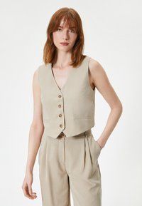 Gilet sans manches beige à encolure en V, cinq boutons marron et deux poches avant. Le matériau semble lisse, associé à un pantalon assorti.
