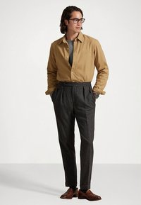 Koszula na guziki w kolorze musztardowym na szarym swetrze, zestawiona z ciemnoszarymi plisowanymi spodniami i brązowymi skórzanymi butami. Prosta bransoletka na nadgarstku.