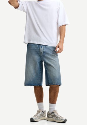 Homme portant un t-shirt blanc oversize, un short en jean bleu clair jusqu'aux genoux, des chaussettes blanches et des baskets beiges, debout devant un fond uni.