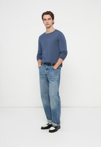 Niebieska koszula z długim rękawem, jasnoniebieskie dżinsy, czarny pasek oraz czarno-białe buty. Model stoi z rękami w kieszeniach na białym tle.