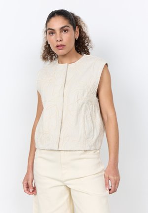 Woman with curly hair wearing a sleeveless beige top with leaf patterns and matching wide-leg pants, standing against a plain white background.