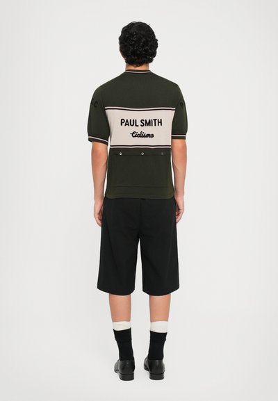 Dunkelgrünes kurzärmliges Hemd mit beigen Streifen, mit "PAUL SMITH"-Logo und "ciclismo"-Text auf dem Rücken. Schwarze Shorts und schwarze Schuhe.