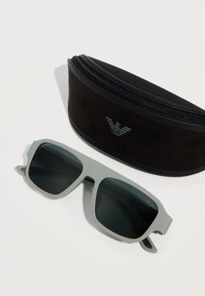 Ορθογώνια γυαλιά ηλίου με γκρι σκελετό και σκούρους φακούς δίπλα σε μια μαύρη θήκη με φερμουάρ, που έχει ασημένιο λογότυπο αετού, πάνω σε λευκή επιφάνεια.