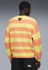 Persoon in een geel en oranje gestreepte trui met een driehoekig logo op de bovenrug, gecombineerd met zwarte broek met een labelpatch.