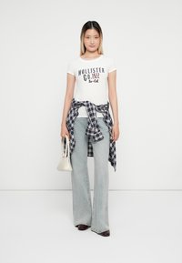 Balta marškinėliai su užrašu "Hollister Co. 1922 So-Cal", šviesiai mėlyni plačios kojos džinsai, languotas marškinis surištas ties liemeniu, ir kreminės spalvos rankinė.