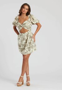Vestido con estampado floral en verde apagado y crema, con mangas abullonadas, un frente retorcido y un corte en la cintura. La modelo lleva sandalias de plataforma.