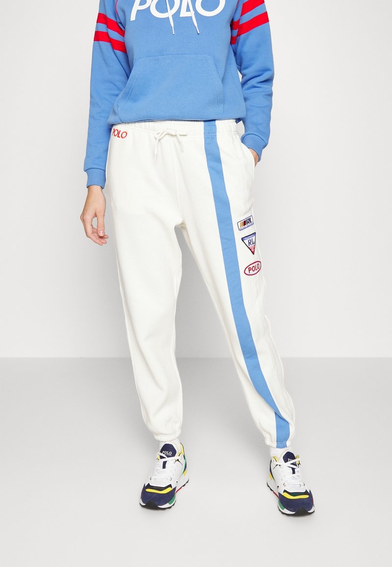 Pantalons de jogging blancs avec une rayure latérale bleue et des patches brodés. Le sweatshirt à capuche bleu comporte des rayures rouges et un logo sur le devant. Baskets multicolores.