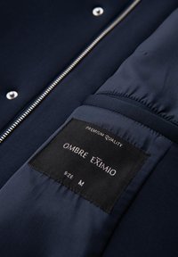 Chaqueta navy con etiqueta de calidad premium, con cremallera y botones de presión. La etiqueta dice "OMBRE EXTIMIO" y talla "M" en texto blanco.