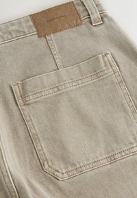 Beige denim pants featuring a back pocket with a stitched, angular design and a brown leather patch labeled "DENIM SUPPLY."