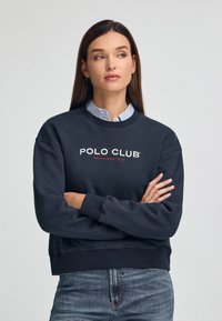 Sudadera de un oscuro color azul marino con un cuello redondo, puños y dobladillo de canalé. Presenta el texto "POLO CLUB" en blanco y rojo, llevada sobre una camisa de cuadros azul.