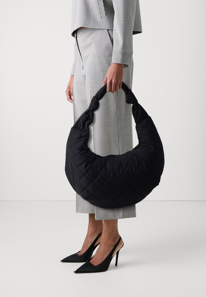 Crna prtljaga s heksagonalnom presvlakom i zakrivljenim oblikom, zalijepena s čvorastom ručkom, izrađena od mekog materijala, u kombinaciji s karičastim odijelom i cipelama s visokom potpeticom.