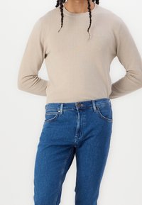 Beige pletený svetr s žebrovaným výstřihem, spojený s modrými džíny s pěti kapsami a zapínáním na knoflík.