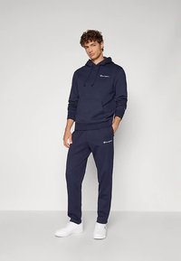 Tumši zils džemperis ar kapuci un auklu, priekšpusē kabata, kopā ar atbilstošām tumši zilām sporta biksēm. Abos ir balts logotipa akcenti. Baltas sporta kurpes.