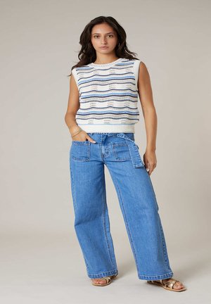 Femme portant un haut en maille sans manches blanc à rayures bleues, un jean bleu pattes larges avec des poches avant, et des sandales dorées, debout avec une main dans la poche.