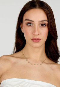 Collana in tono dorato con sette gemme blu rotonde, equidistanti, esposta su un collo nudo e un top senza spalline.