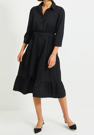Robe noire à manches trois-quarts, avec un devant à boutons, une ceinture à nouer à la taille et un ourlet à volants en couches. Tissu lisse, design longueur cheville.