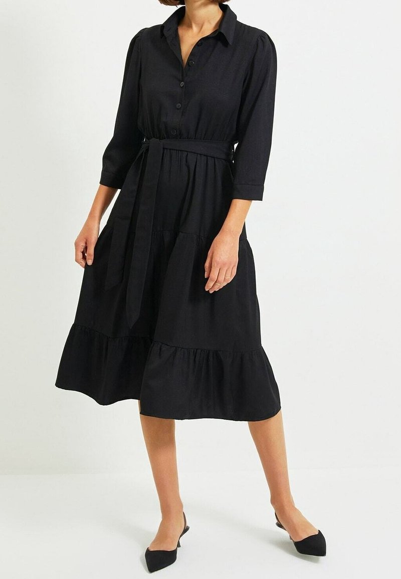 Vestito nero a camicia con maniche a tre quarti, chiusura a bottoni sul davanti, cintura in vita e orlo a balze stratificate. Tessuto morbido, design lungo fino alla caviglia.