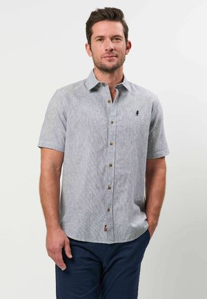 Man draagt een lichtgrijs overhemd met korte mouwen en knopen, gecombineerd met een donkerblauwe broek. Hij staat met één hand in zijn zak tegen een effen achtergrond.