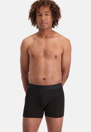 Man met krullend haar die zwarte boxers draagt, staand tegen een effen witte achtergrond, handen achter de rug, voorwaarts gericht.