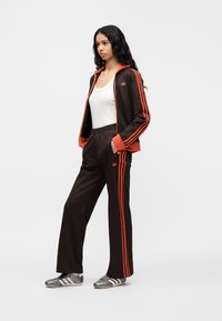 Καφέ φόρμα με πορτοκαλί ρίγες, που περιλαμβάνει μπουφάν με φερμουάρ και παντελόνια με λάστιχο. Άσπρο αμάνικο τοπ από μέσα, γκρι αθλητικά παπούτσια ολοκληρώνουν την εμφάνιση.