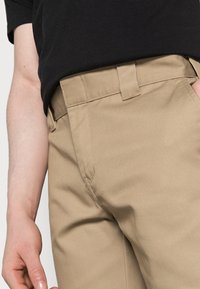 Pantaloni kaki cu o textură netedă, având o croială dreaptă, buzunare laterale și un tiv cusut pentru o potrivire sigură. Un top negru este parțial vizibil.