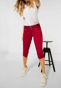 Pantaloni rossi cropped realizzati in morbido cotone, con un design aderente, due tasche frontali e chiusura a bottone. Indossati con una camicia bianca e sneakers.