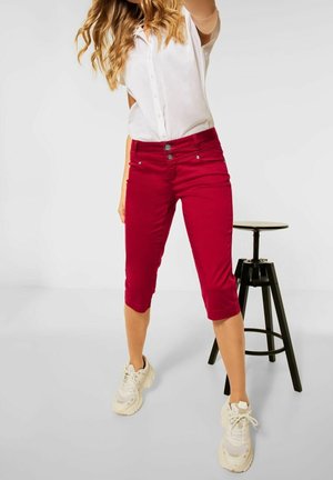 Pantalones cortos rojos de algodón suave, con un diseño ajustado, dos bolsillos frontales y cierre de botón. Llevados con una camisa blanca y zapatillas.
