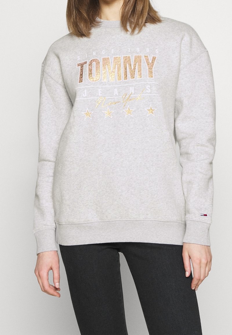 Sudadera gris claro con cuello de barco. Presenta "TOMMY JEANS" en impresión de purpurina dorada, cinco estrellas doradas debajo y puños y dobladillo de canalé.