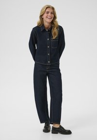 Conjunto de chaqueta de mezclilla oscura y pantalones de pierna ancha, con costuras en contraste, cierre de botón y un bolsillo en el pecho. Combinado con calzado negro.
