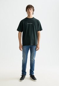 Camiseta de manga corta de color verde oscuro con texto de marca en blanco, combinada con vaqueros azul claro y zapatillas negras. Diseño sencillo con un ajuste relajado.