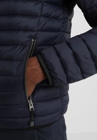 Marineblauer Daunenjacke mit gestepptem Design, ausgestattet mit einer Reißverschlusstasche auf der linken Seite und gerippten Bündchen für eine enganliegende Passform.