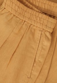 Gros plan sur un pantalon en tissu marron clair avec une ceinture élastique et une poche latérale visible.