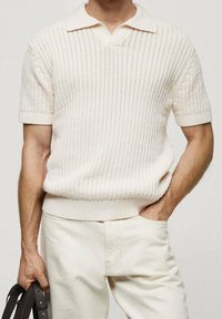 Hombre con una camisa polo de punto de manga corta color crema y pantalones color crema, sosteniendo un cinturón de cuero marrón oscuro en una mano.