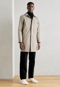 Beige vedenkestävä trench-kot, jossa on kaulus ja yksittäinen napitus, yhdistettynä mustiin housuihin ja valkoisiin tennareihin puisella lattia- taustalla.