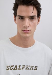 Młody mężczyzna z ciemnymi włosami, ubrany w białą koszulę z napisem "SCALPERS" w postarzanych brązowych literach.