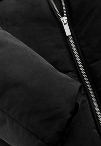 Veste matelassée noire avec une texture de tissu lisse, un rembourrage épais et une fermeture éclair argentée sur le devant. Une manche arrondie est visible.
