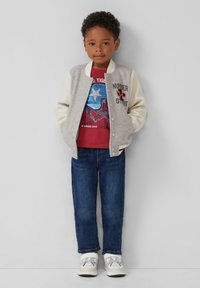 Graue und cremefarbene Varsity-Jacke mit Druckknöpfen, rotes T-Shirt mit grafischem Design eines silbernen Bären, blaue Jeans und weiße Sneakers.
