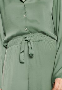 Grön, silkeslen tvådelad outfit med en knäppning-up topp med långa ärmar och ett elastiskt midjeband med en knuten rosett vid midjan.