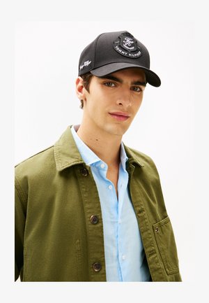 Ung mand iført sort Tommy Hilfiger kasket, lyseblå skjorte med knapper og olivengrøn jakke med store knapper og brystlomme.