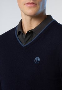 Dunkelblauer Pullover mit V-Ausschnitt, der über einen kontrastierenden hellblauen Saum und ein dunkelgraues Hemd darunter verfügt. Kleines Logo auf dem Pullover.