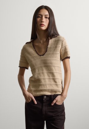 Frau mit langem dunklem Haar trägt ein beiges, gehäkeltes Kurzarmoberteil mit dunklem Rand und schwarze Hosen, steht mit den Händen in den Taschen vor einfarbigem Hintergrund.