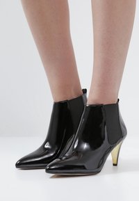 Bottines en cuir verni noir avec des orteils pointus, des panneaux latéraux élastiques et des talons aiguille dorés. Texture lisse avec une finition brillante.