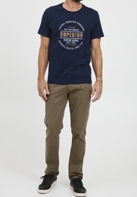 Granatowy bawełniany t-shirt z okrągłym nadrukiem tekstowym w kolorach brązowym i białym, zestawiony z jasnobrązowymi spodniami i czarnymi trampkami z białymi podeszwami.